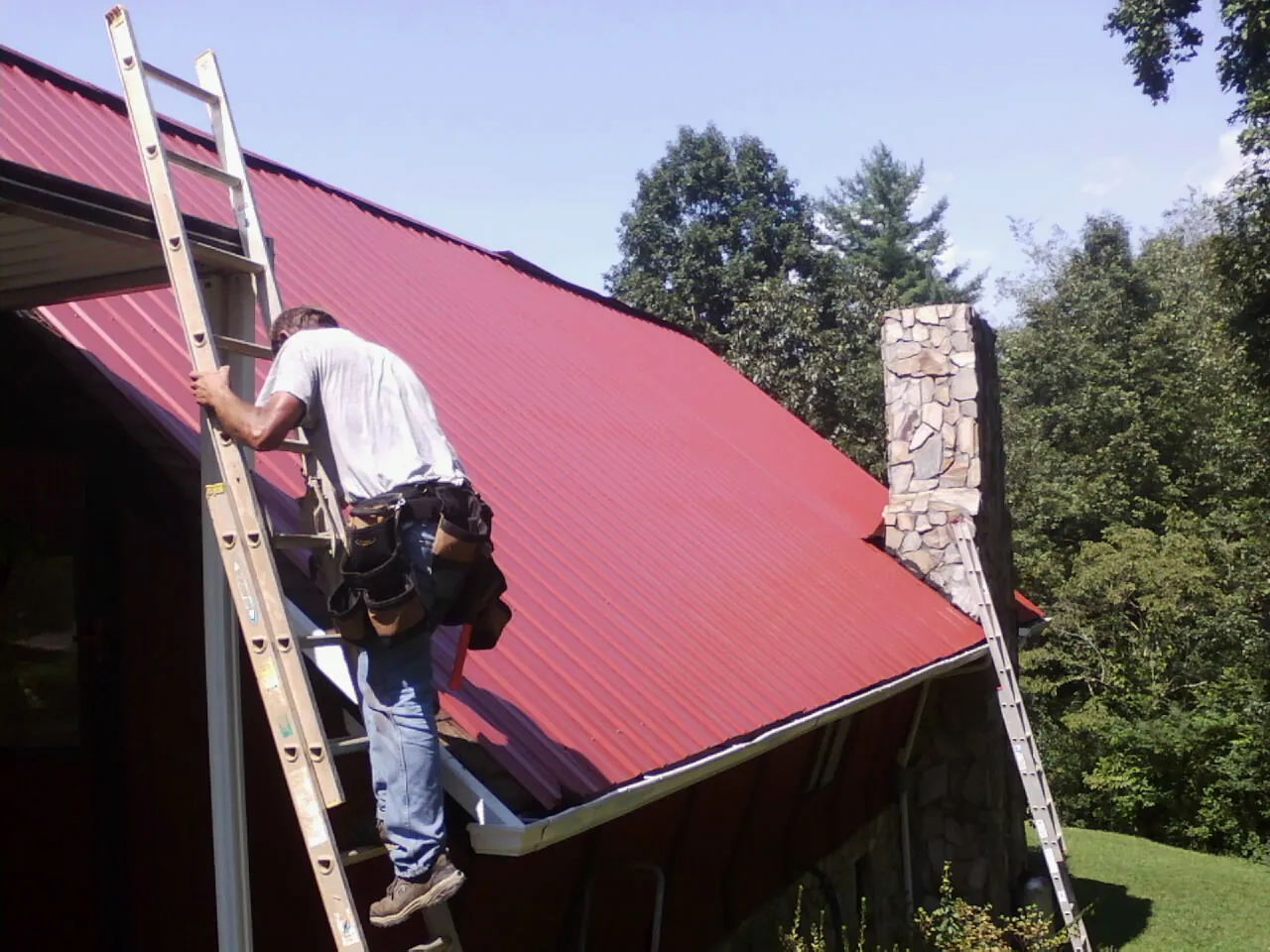 Roofer on a ladder beside a red metal roof and stone chimney — Soffit & Fascia Repair by Roofing Brothers in Stewartstown